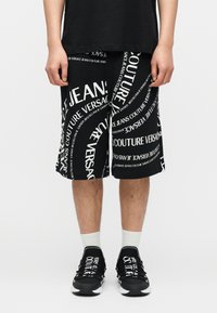 Άτομο που φοράει μαύρο σορτς με λευκό μοτίβο κειμένου Versace Jeans Couture, λευκές κάλτσες και ταιριαστά μαύρα αθλητικά παπούτσια με λευκό κείμενο.