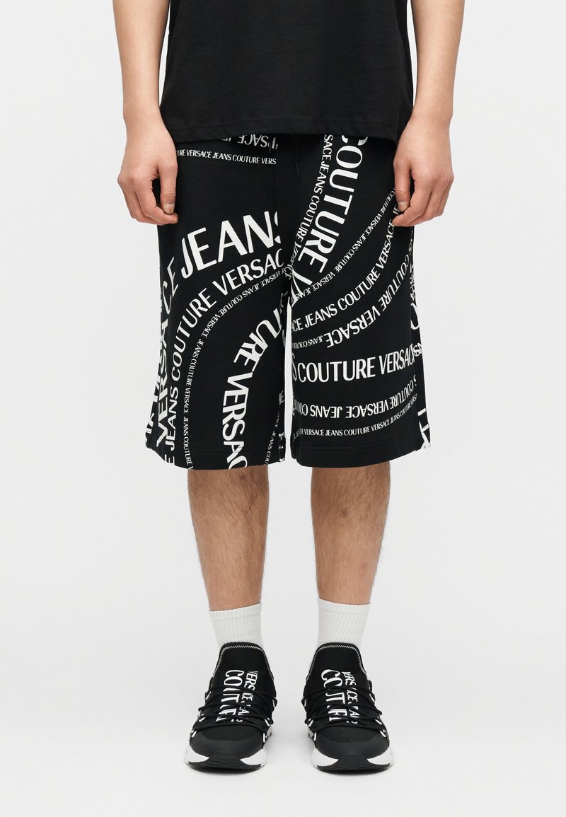 Άτομο που φοράει μαύρο σορτς με λευκό μοτίβο κειμένου Versace Jeans Couture, λευκές κάλτσες και ταιριαστά μαύρα αθλητικά παπούτσια με λευκό κείμενο.