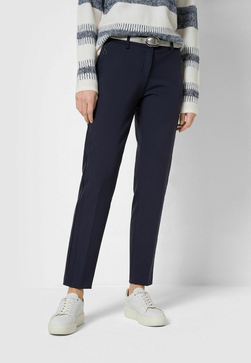 Pantaloni navy con vestibilità slim e frontale piatto, dotati di passanti per cintura e tasca con zip, abbinati a scarpe da ginnastica bianche e a un maglione a righe.