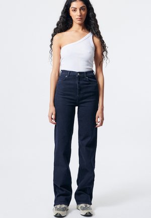 Jeune femme aux longs cheveux bouclés portant un haut blanc à une épaule, un jean sombre taille haute et des baskets, debout devant un fond clair.