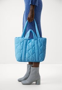 Gequiltede blauwe draagtas met twee handvatten; textuur met gestikte patronen. Gepaard met grijze haklaarzen.