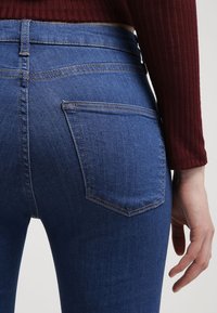 Blå denimjeans med hög midja och en mjuk textur, med dubbla bakfickor och kontrasterande sömmar. Figurformad siluett och ingen synlig hårdvara.