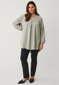 Lichtgroene blouse met lange mouwen en een V-hals, gemaakt van zachte, gestructureerde stof. Gecombineerd met zwarte slim-fit broek en schoenen met strikdetail.