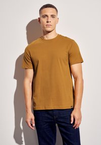 Tričko s krátkým rukávem a kulatým výstřihem v teplém hnědém odstínu. Vyrobeno z hladkého materiálu, v kombinaci s tmavě modrými džínami. Visačka viditelná na lemu.