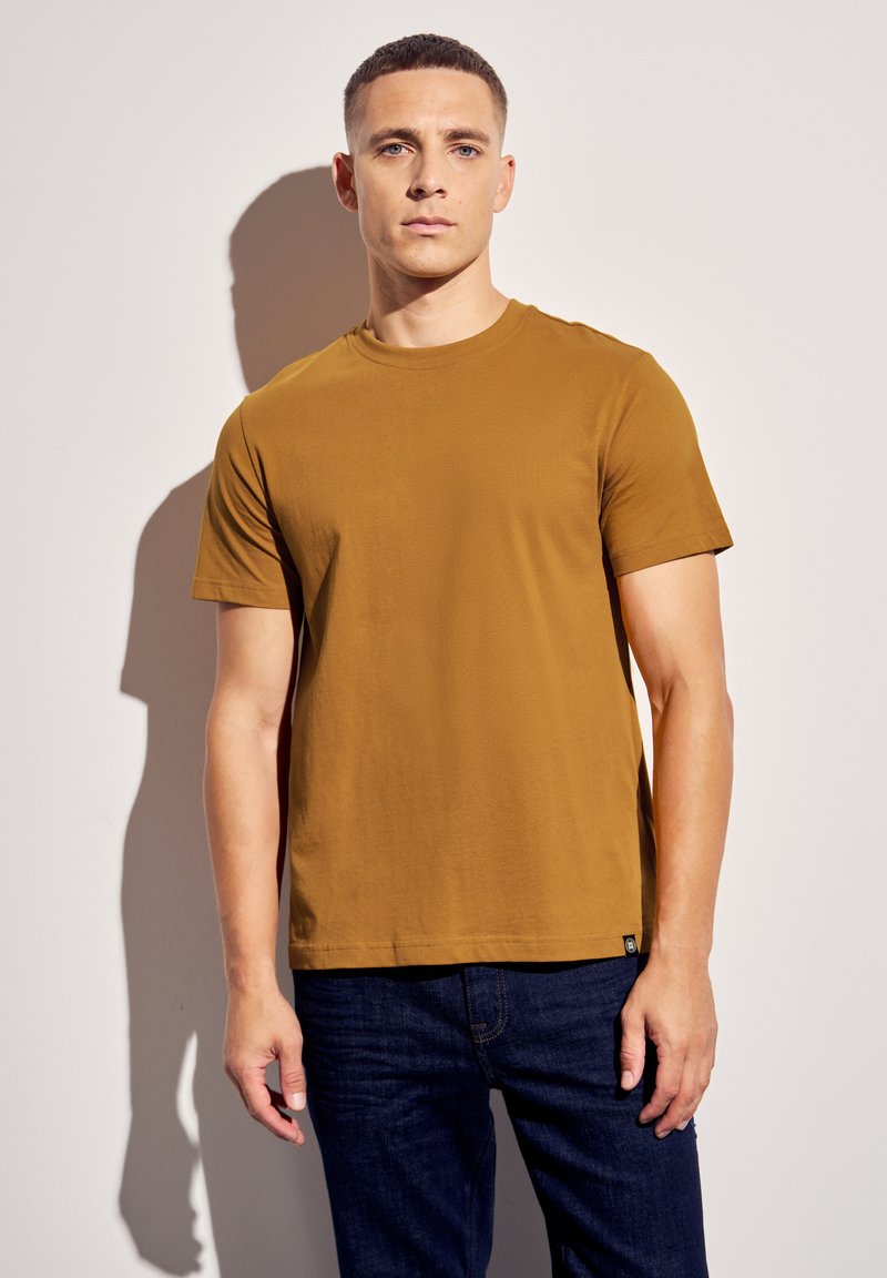 Tričko s krátkým rukávem a kulatým výstřihem v teplém hnědém odstínu. Vyrobeno z hladkého materiálu, v kombinaci s tmavě modrými džínami. Visačka viditelná na lemu.
