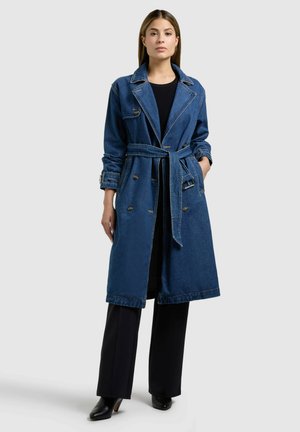 Vrouw die een blauw denim trenchcoat tot op de knie draagt met een ceintuur in de taille, een zwarte top, zwarte wijde broekspijpen en zwarte laarzen met hak, op een effen achtergrond.