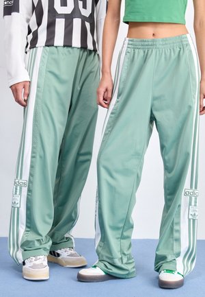 Twee personen dragen bijpassende losse mintgroene Adidas trainingsbroeken met witte zijstrepen en sneakers, staande op een blauwe vloer.