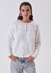 Cardigan bianco lavorato a maglia con applicazioni floreali testurizzate, scollatura rotonda, chiusure a bottoni e polsini e orlo a coste. Abbinato a jeans grigi sbiaditi.