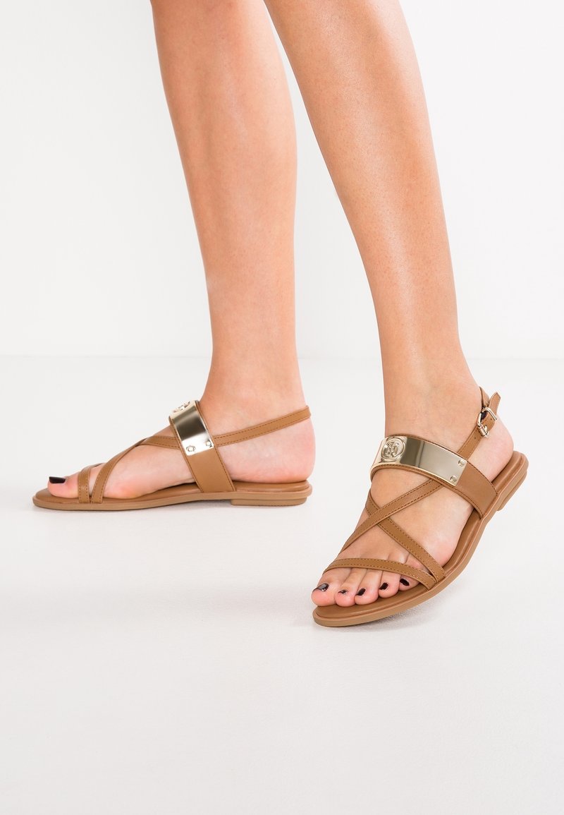 Jambes portant des sandales plates couleur beige avec un détail de sangle métallique dorée, ongles de pieds peints en noir, debout sur un fond blanc.