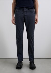 Dark blue cotton chinos with a slim fit, feature front pockets, a button closure, and a smooth texture. Black shoes are worn.