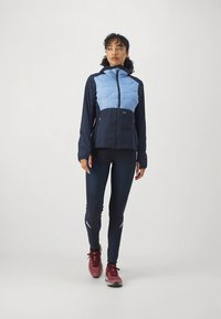 Svijetloplava i tamnoplava podstavljena jakna s patentnim zatvaračem sprijeda, uparena s tamnim tajicama i crvenim tenisicama. Ima reflektirajuće detalje na nogama.