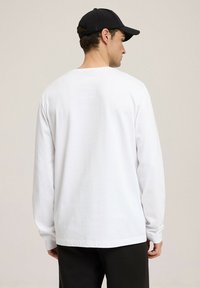 Biała koszula z długim rękawem i okrągłym dekoltem, wykonana z miękkiej bawełny, o luźnym kroju i prostym wykończeniu. Noszona z czarną czapką i ciemnymi spodniami.