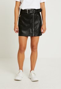 Falda negra de piel sintética con cintura alta, frente con cremallera y presillas para cinturón. Combinada con zapatillas blancas. Textura suave y diseño ajustado.