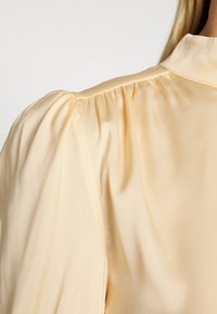 Blouse jaune pâle à texture satinée, avec des manches bouffantes froncées et un col à boutons. Tissu lisse avec un léger éclat.