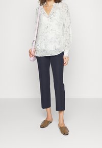 Kvinna i en genomskinlig vit blommig blus, avkapade marinblå byxor, bruna slip-on skor och bär en ljusrosa axelväska.