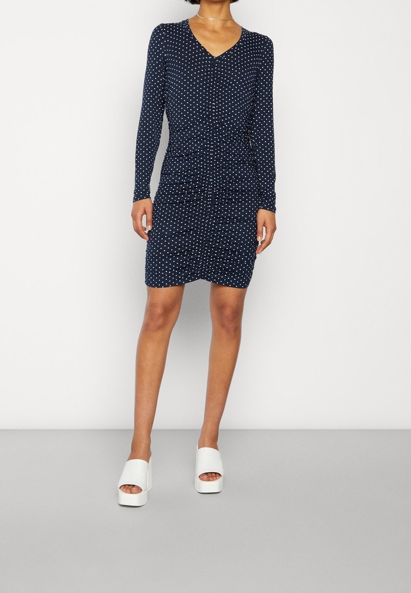 Femme vêtue d’une robe ajustée bleu marine à pois avec manches longues et col en V, associée à des sandales blanches compensées.