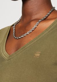 Oliwkowa koszulka w serek z krótkimi rękawami i prążkowaną teksturą, z dyskretnym logo na piersiach oraz plecionym srebrnym naszyjnikiem.