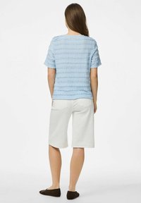 Gaiši zils teksturēts tops ar horizontālām svītrām, apvienots ar baltām līdz ceļiem īsām biksēm un brūniem slēgtiem apaviem.