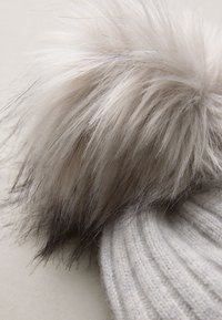 Siva pletena kapa z velikim, puhastim cofkom na vrhu. Cofek ima prelivanje od svetlo do temno sivih odtenkov s finimi nitmi.