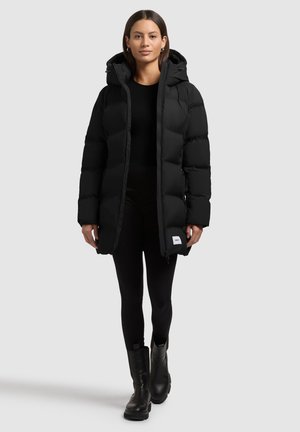 Chaqueta de plumas negra con capucha, con cremallera en la parte delantera, bolsillos laterales y un acabado texturizado. Llevada sobre leggings negros y botas.