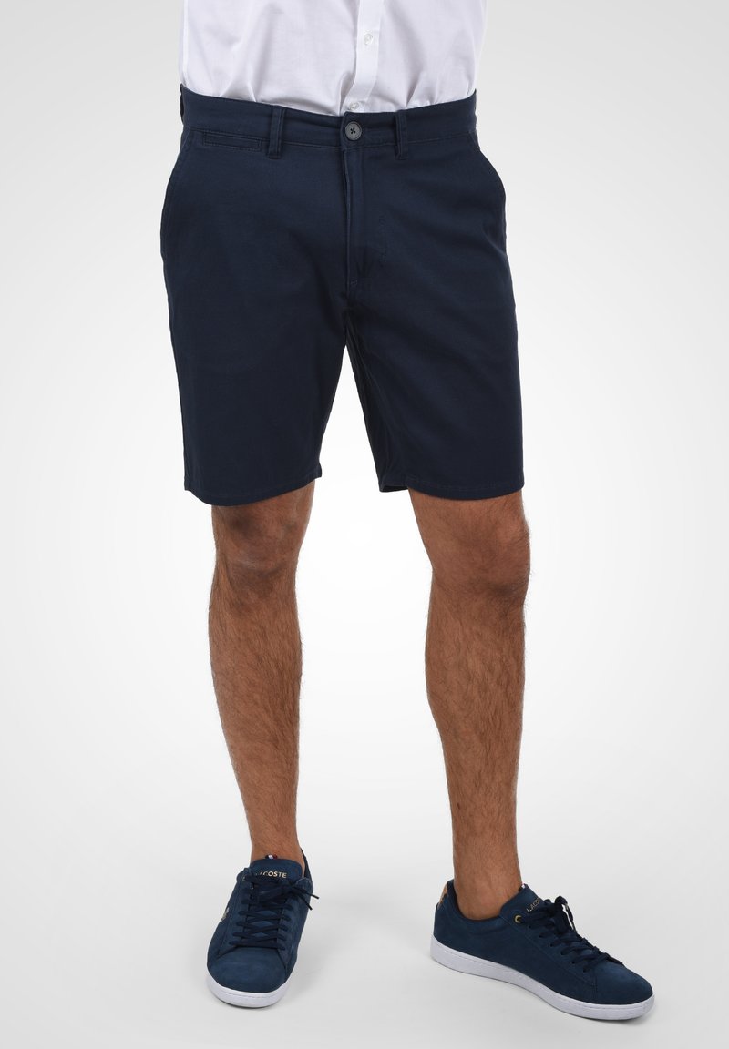 Shorts en coton bleu marine avec fermeture à bouton, poches avant et coupe droite. Portés avec des baskets assorties bleu foncé.