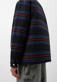 Wolljacke mit schwarzem Grund und horizontalen roten, grünen und blauen Streifen. Das Design umfasst goldene Knöpfe an den Ärmeln und eine legere Passform.