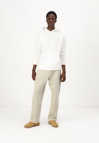 Hanorac alb cu șnururi, buzunar tip kangur și detaliu cu logo. Pantaloni cargo bej cu accente de cusătură. Sneakers maro cu sistem de �șiret.