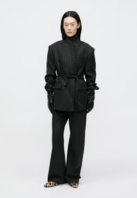 Fato preto riscado com um casaco de lapela dupla e cinto, calças de perna larga e luvas justas pretas. Sapatos de estampado de leopardo.