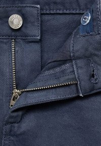 Razvijene tamnoplave traperice s metalnim zatvaračem srebrne boje, metalnom dugmadi označenom "MNG DENIM" i malim prozirnim plastičnim dugmadi.