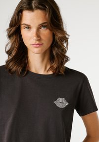 Νεαρή γυναίκα με καστανό μαλλί σε μήκος ώμου και κυματιστό στυλ, φορώντας ένα μαύρο t-shirt με λογότυπο "Pepe Jeans London" σε σχήμα χειλιών στο στήθος.