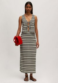 Schwarz-weißer gestreifter ärmelloser Kleid mit einem Frontbindedetail, ergänzt durch eine rote rechteckige Handtasche und flache schwarze Sandalen.