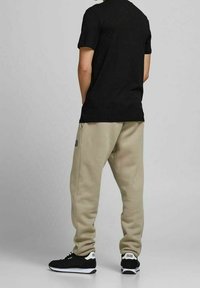 Béžové tepláky s elastickými manžetami, kombinované s černým tričkem s krátkým rukávem a černými teniskami s bílými akcenty, viděné ze zadu.