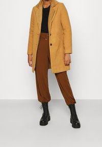 Manteau en mélange de laine moutarde avec un col à revers et deux boutons, porté sur un haut noir à côtes et un pantalon marron fuselé. Bottes noires à lacets.