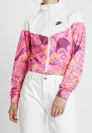 Vrouw draagt een witte Nike-jas met ritssluiting, versierd met roze, paarse en gele bloemmotieven op de mouwen en borst, gecombineerd met een witte broek.