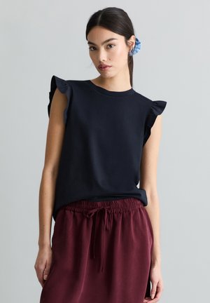 Junge Frau mit dunklem Haar, zurückgebunden mit einem blauen Haargummi, trägt ein schwarzes ärmelloses Oberteil mit Rüschenschultern und burgunderrote Hose mit Kordelzug.