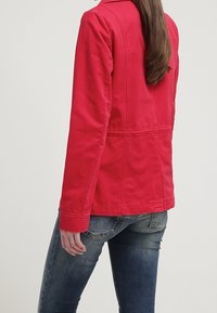 Chaqueta de algodón roja con un ajuste entallado, manga larga y cuello. Presenta detalles de costuras y textura suave. Se lleva con vaqueros azules.