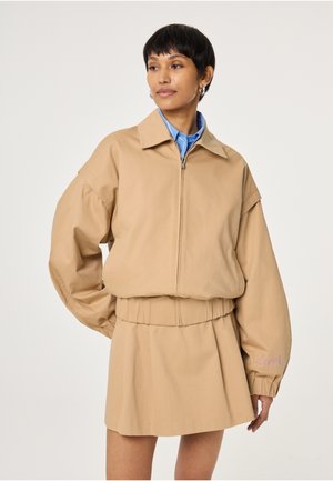 Vrouw draagt een beige jas met ritssluiting en elastische cuffs en bijpassende beige rok, staand tegen een effen lichte achtergrond.