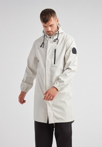 Impermeabile leggero beige con cappuccio, polsini elasticizzati, tasca con zip frontale e una toppa nera con il logo sulla manica. Texture liscia, lunghezza fino al ginocchio.
