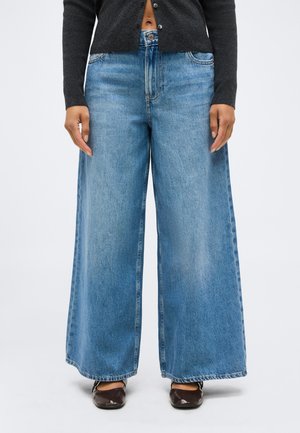 Personne portant un jean léger bleu clair, ample et à jambes larges, un haut court à manches longues foncé, et des chaussures plates marron foncé, debout devant un fond blanc.