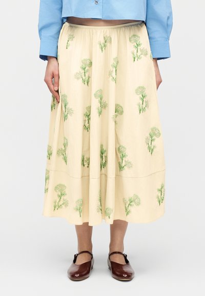 Μπεζ midi φούστα με πράσινη κεντητή λουλουδάτη διακόσμηση, φορεμένη με καφέ παπούτσια Mary Jane και ελαφρύ μπλε κοντό πουκάμισο, εμφανίζονται από τη μέση μέχρι τα πόδια.