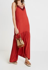 Czerwona maxi sukienka na ramiączkach spaghetti z koronkowymi akcentami. Lekka, zwiewna tkanina, asymetryczny dół. Zestawiona z teksturowaną torebką i sandałami.