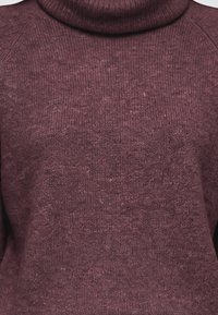Gros plan d’un pull à col roulé en maille douce de couleur mauve foncé avec une texture côtelée subtile sur le cou et le haut de la poitrine.