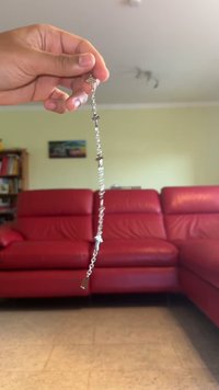 Stříbrný řetízek s několika malými kříži, texturovanými články a obdélníkovým přívěskem visícím na konci, držený v ruce před červenou pohovkou.