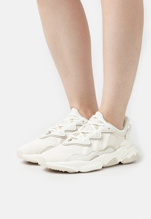 Zapatillas deportivas con una parte superior texturizada de color blanco roto, con cordones y una suela acolchada. Punta redondeada y detalles de marca sutiles.