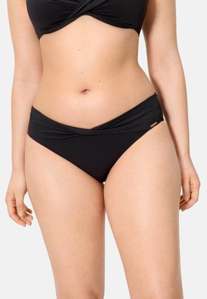 Mujer con braguita de bikini negra con detalle de cintura torcida, de pie contra un fondo claro y liso.