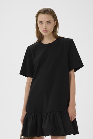 Jeune femme aux cheveux châtain clair portant une robe noire à manches courtes avec un ourlet volanté, debout devant un fond clair uni.