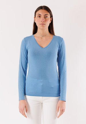 Donna con lunghi capelli scuri che indossa una maglia blu a maniche lunghe con scollo a V e pantaloni bianchi, in piedi davanti a uno sfondo chiaro e uniforme.
