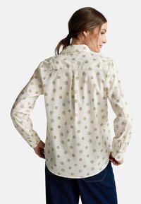 Cremefarbene Bluse mit Rosenmuster in Gold und Pink, langen Ärmeln, Knopfkragen und geschwungenem Saum. Hergestellt aus leichtem Stoff.