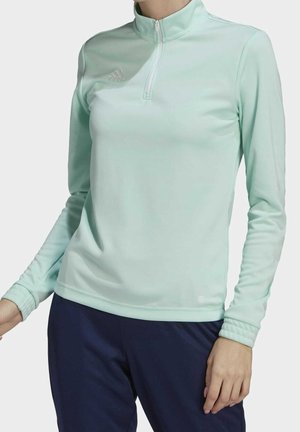 Femme portant un haut zippé à manches longues bleu clair et un pantalon bleu foncé sur fond blanc uni.