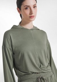 Długa bluza z kapturem w kolorze oliwkowej zieleni, wykonana z miękkiego materiału, z przednim wiązaniem i kapturem. Gładka tekstura, luźny krój.